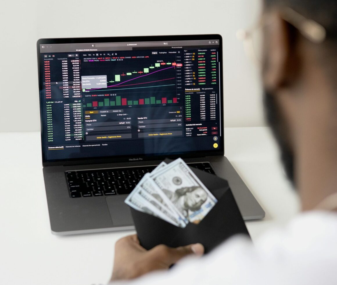 A person examines stock market data on a laptop while holding dollar bills, showcasing finance and trading concepts.
